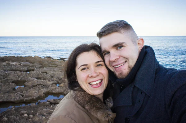 Retrato de jovem casal bonito tirando foto selfie em um smartphone com mar e céu escuro nublado no fundo. Temporada fria e conceito de viagem . — Fotografia de Stock