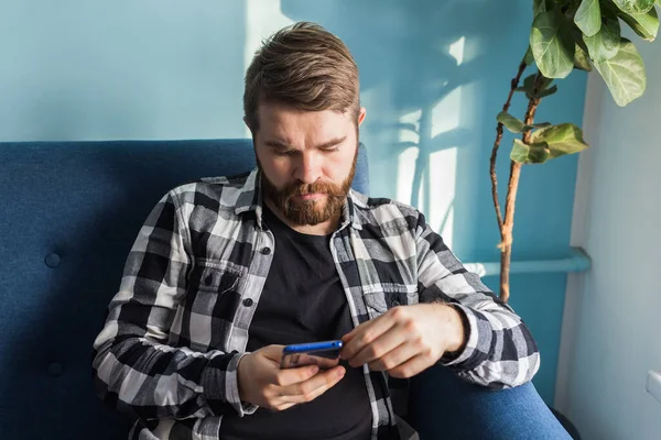 Tecnologías y concepto de ocio - Hombre alegre sentado en el sofá usando su teléfono inteligente en casa en la sala de estar . — Foto de Stock