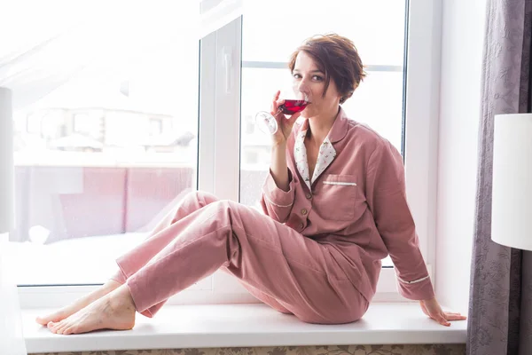 Mulher bonita usa pijama com um copo de vinho tinto senta-se no peitoril da janela perto da bela janela em casa. Fica em casa. Quarentena e coronavírus covid-19 pandemia . — Fotografia de Stock
