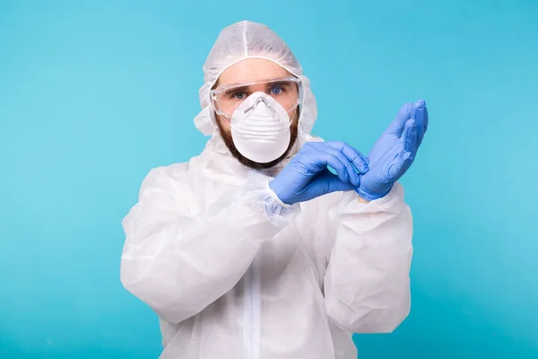 Людина в білому костюмі для дезактивації одягає медичні рукавички, ізольовані на синьому тлі. Coronavirus, covid-19 and pandemic concept. — стокове фото