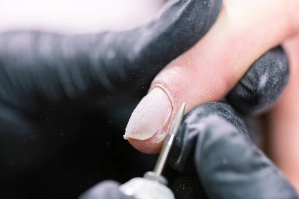 Крупный план профессиональных косметологов, работающих с электрической дрелью на ногтях клиента. Процедура нанесения искусственных ногтей. Руки маникюрши с инструментом. Маникюрша в перчатках удаляет кутикулу — стоковое фото