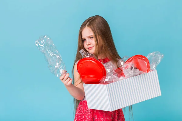 Niña impactada mira con los ojos abiertos y la expresión preocupada, la celebración de la caja con varios residuos de plástico en el fondo azul . —  Fotos de Stock