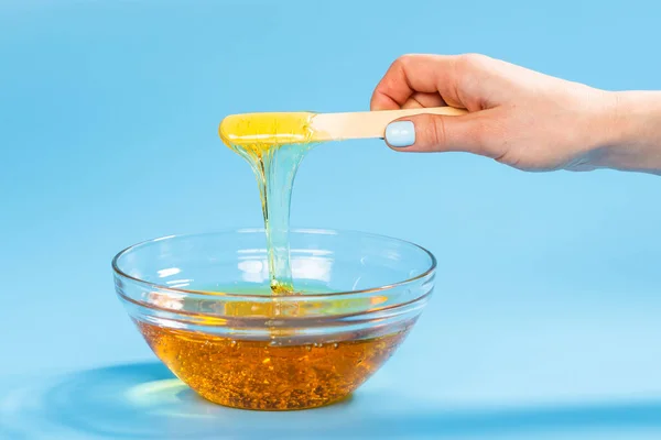 Liquid yellow wax or sugar paste for depilation drains from the stick on blue background. The concept of depilation, waxing, sugaring smooth skin without hair.