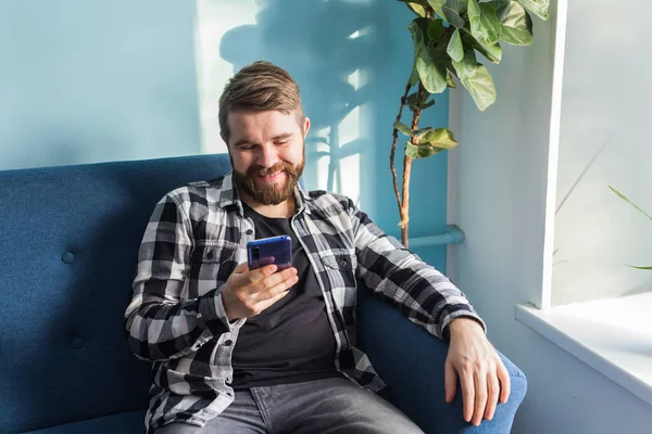 Hombre jugando juegos en línea con un teléfono inteligente sentado en un sofá en casa. — Foto de Stock