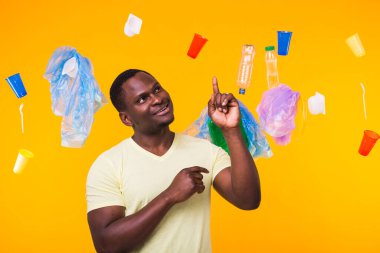 Dünya Çevre Günü, plastik geri dönüşüm sorunu ve çevresel felaket konsepti - sarı arka planda çöplere bakan afro-amerikan erkeği.