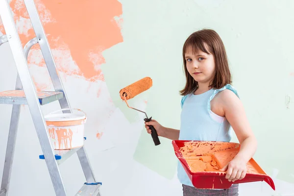 Reparación en el apartamento. La niña feliz pinta la pared con pintura naranja. Concepto de renovación y redecoración . — Foto de Stock