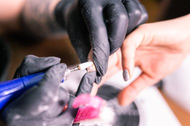 Kadın usta kuaförde manikür yaparken ojeyi çıkarmak için elektrikli makine kullanıyor. Yakın çekim donanım manikürü. El bakımı kavramı. Kadın manikürcü, bir değirmen kesicisiyle tırnaklarını temizliyor.