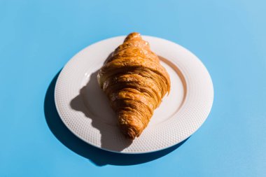 Fırın ürünleri beyaz tabakta pişmiş kruvasan. Mavi arkaplan, en üstteki yakın plan. Pop sanat tarzı. Yaz gölgeleri. Lezzetli ve yemek konsepti.