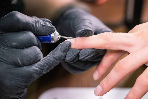 Κοντινά χέρια επαγγελματία αισθητικός που εργάζονται με ηλεκτρικό τρυπάνι στα νύχια του πελάτη. Διαδικασία εφαρμογής τεχνητών νυχιών. Χέρια μανικιουρίστα με εργαλείο. Μανικιουρίστας στα γάντια αφαιρεί την επιδερμίδα — Φωτογραφία Αρχείου