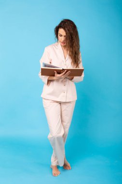 Pijamalı gülümseyen genç kız, mavi arka plan stüdyo portresi üzerine bir kitap okuyor. Rahatla iyi ruh hali yaşam tarzı konsepti.