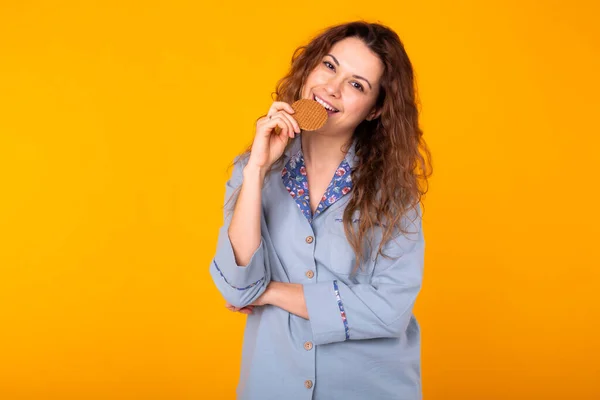 Joyful woman wears fashion pajama going to eat delicious cookie on yellow background with copy space. Morning, breakfast and home wear concept.