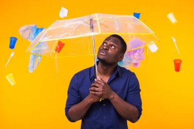 Dünya Çevre Günü, plastik geri dönüşüm sorunu ve çevresel felaket konsepti - sarı arka planda çöplere bakan afro-amerikan erkeği.