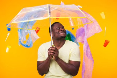 Dünya Çevre Günü, plastik geri dönüşüm sorunu ve çevresel felaket konsepti - sarı arka planda çöplere bakan afro-amerikan erkeği.