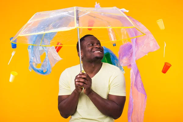 Dünya Çevre Günü, plastik geri dönüşüm sorunu ve çevresel felaket konsepti - sarı arka planda çöplere bakan afro-amerikan erkeği.