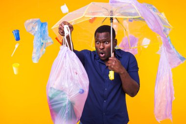 Plastik geri dönüşüm sorunu, ekoloji ve çevre felaketi kavramı. Sarı arka planda çöplerle yaşayan Afro-Amerikan erkeği şaşırttı. Ekoloji felaketinden endişeleniyor.