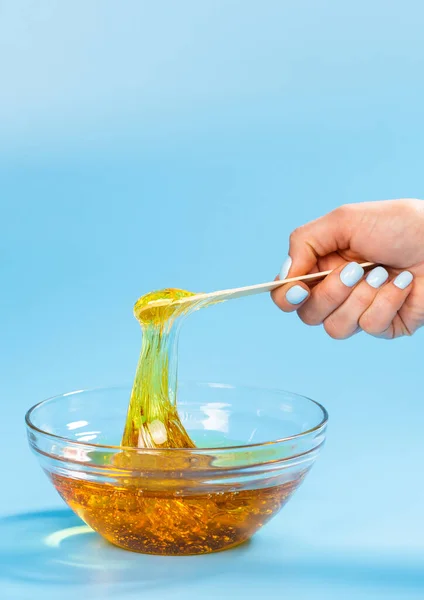 Kvinnliga händer som håller flytande gul pasta för socker på blå bakgrund. Depilering och hårborttagning koncept. — Stockfoto