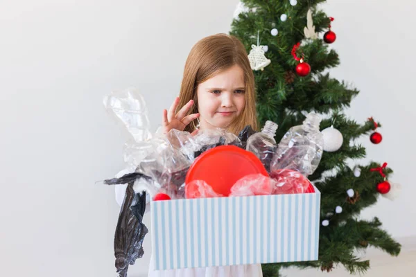 Schockiertes kleines Mädchen blickt mit geöffneten Augen und besorgtem Gesichtsausdruck und hält Schachtel mit verschiedenen Plastikabfällen über dem Christbaumhintergrund. — Stockfoto