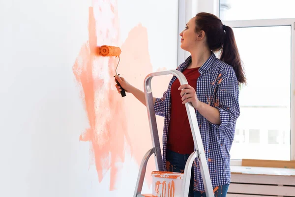 Happy young woman painting interior wall with paint roller in new house. A woman with roller applying paint on a wall.