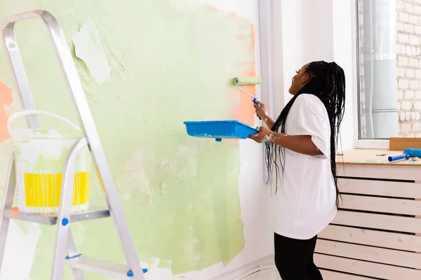 O femeie afro-americană zâmbitoare care pictează peretele interior al unei case noi. Redecorare, renovare, reparații apartamente și concept de băuturi răcoritoare . — Fotografie, imagine de stoc