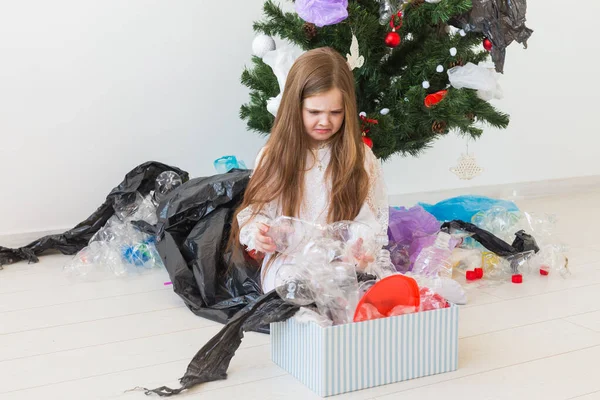 Niña impactada mira con los ojos abiertos y expresión preocupada, caja de espera con varios residuos de plástico sobre el fondo del árbol de Navidad . — Foto de Stock