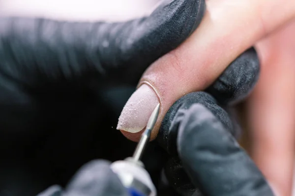 Primer plano profesional esteticista manos que trabajan con taladro eléctrico en las uñas del cliente. Procedimiento de aplicación de uñas artificiales. Manos de manicura con herramienta. La manicura en guantes elimina la cutícula —  Fotos de Stock