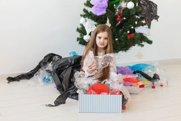 Niña impactada mira con los ojos abiertos y expresión preocupada, caja de espera con varios residuos de plástico sobre el fondo del árbol de Navidad . — Foto de Stock