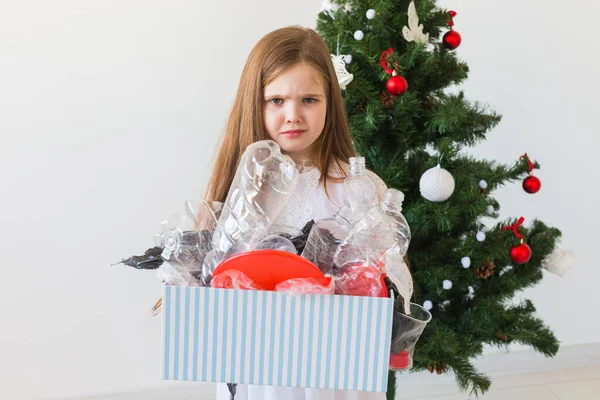 Schockiertes kleines Mädchen blickt mit geöffneten Augen und besorgtem Gesichtsausdruck und hält Schachtel mit verschiedenen Plastikabfällen über dem Christbaumhintergrund. — Stockfoto