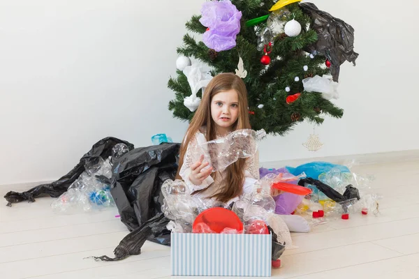 Niña impactada mira con los ojos abiertos y expresión preocupada, caja de espera con varios residuos de plástico sobre el fondo del árbol de Navidad . — Foto de Stock