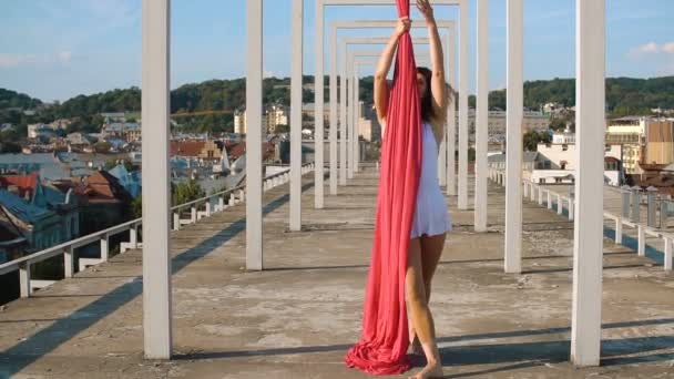 Bela dançarina em seda aérea. Jovem mulher sexy realiza acrobacias acrobáticas no telhado contra o céu com nuvens — Vídeo de Stock