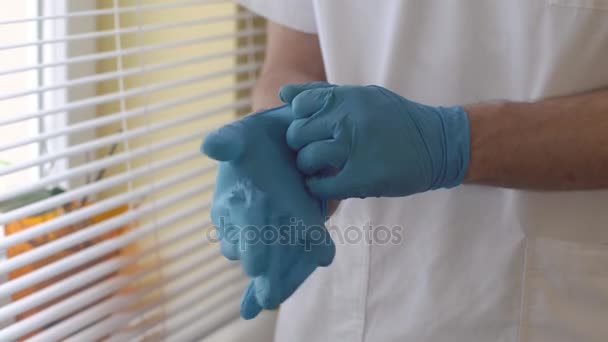 Doctor de cerca poniéndose un guante de látex azul esterilizado. Médico varón de pie en el consultorio médico cerca de la ventana . — Vídeos de Stock