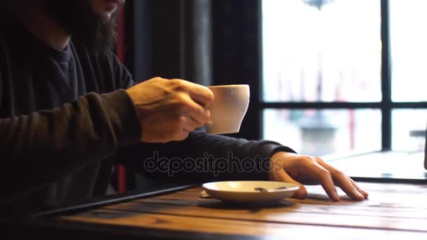 Joven bebiendo café junto a la ventana del café. Un hombre con barba . — Vídeos de Stock