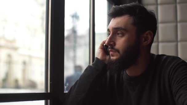 Jonge man praten op mobiele telefoon in de buurt van het venster in het café. Een man in een slecht humeur. Close-up, 4k. — Stockvideo