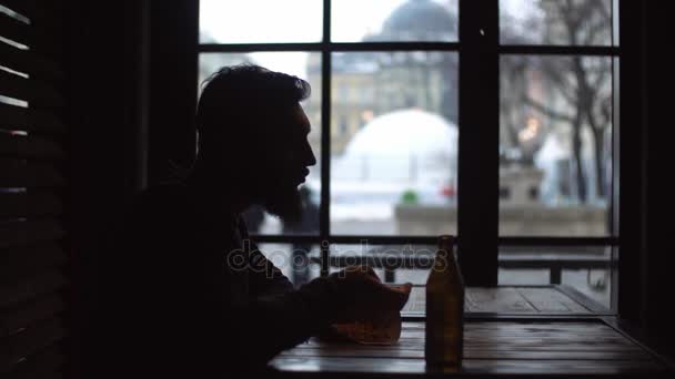 Sylwetka młodego człowieka, który jest picie piwa i jedzenia przekąski w barze przy oknie. — Wideo stockowe