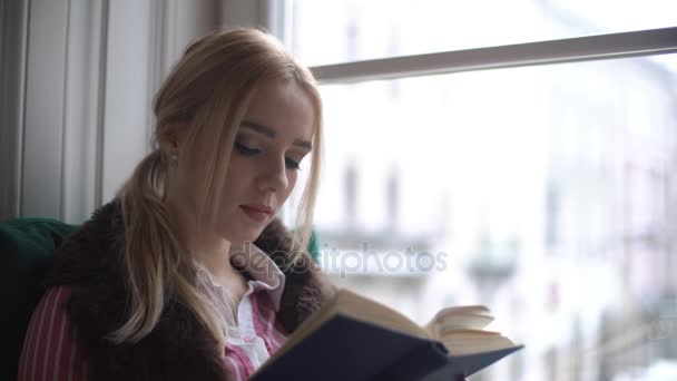 Hermosa joven sentada en el alféizar de la ventana, leyendo un libro y mirando por la ventana — Vídeos de Stock