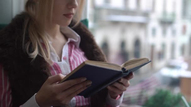 Hermosa joven sentada en el alféizar de la ventana, leyendo un libro y mirando por la ventana — Vídeos de Stock