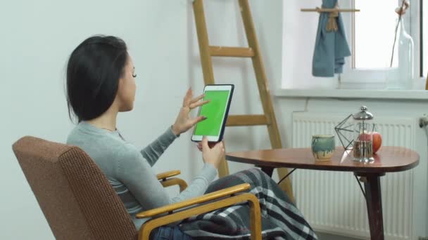 Όμορφο κορίτσι χρησιμοποιώντας tablet με πράσινη οθόνη, κάθεται σε μια άνετη καρέκλα στο σπίτι — Αρχείο Βίντεο