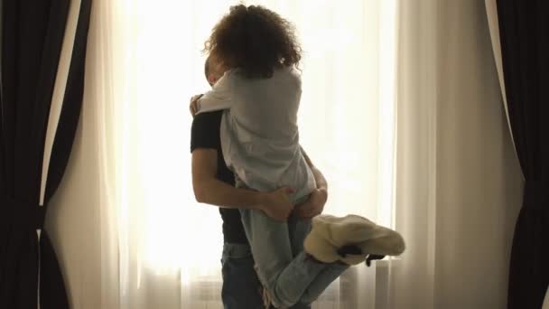 Young beautiful couple fooling around at home by the window. The young man takes the girl up in his arms and whirls with her — Stock Video