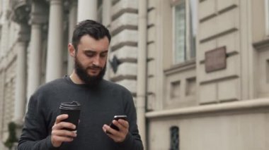 Mutlu sakal genç adam manifatura smartphone ve içme kahve sokakta günbatımında. O sokak, hareket kamerayı yürüyor
