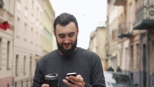 Happy beard young man texting on smartphone and drinking coffee in the street at sunset. He walks down the street, motion camera — Stock Video