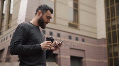 Mutlu sakal genç adam manifatura smartphone ve kahve sokakta günbatımında içme.