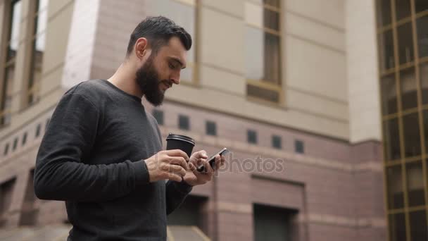 Mutlu sakal genç adam manifatura smartphone ve kahve sokakta günbatımında içme. — Stok video