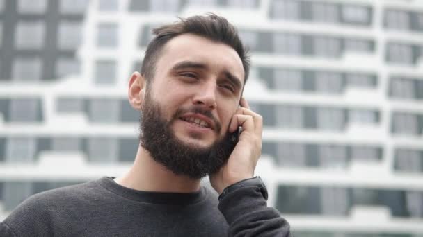 Молодой человек с бородой разговаривает по телефону возле высоток — стоковое видео