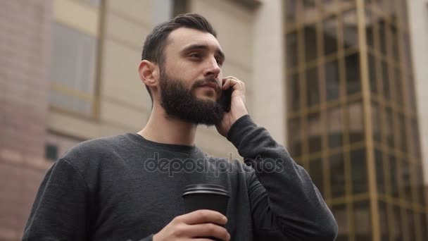 Feliz barba joven hablando en el teléfono inteligente y tomando café en la calle al atardecer — Vídeos de Stock