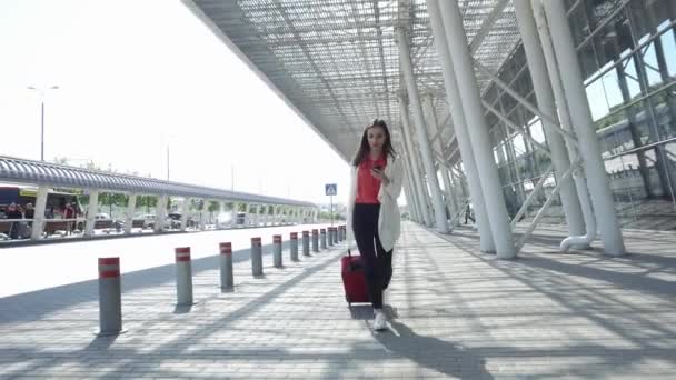 Жінка в червоній сорочці говорить по телефону, ходячи з валізою — стокове відео