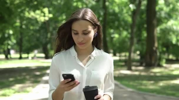 Vrouw drinkt koffie en controleert haar telefoon in het park — Stockvideo