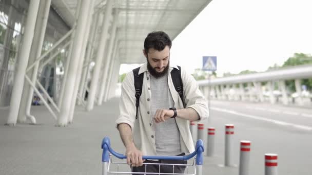 Smiling bearded man walks with a rucksack and checks his smart watch — Stock Video