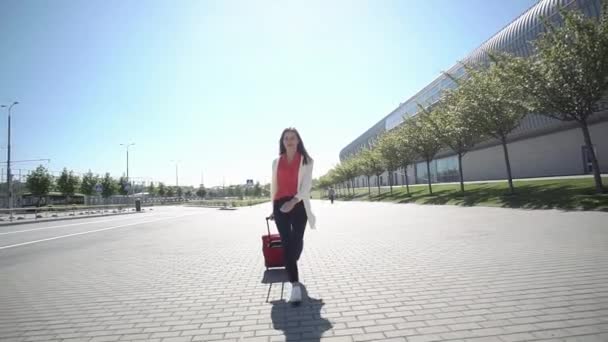 Mujer bonita con camisa roja camina con la maleta afuera en un día soleado — Vídeo de stock