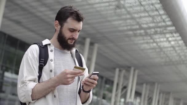 Усміхнений бородатий чоловік ходить з рюкзаком і перевіряє свою кредитну картку — стокове відео