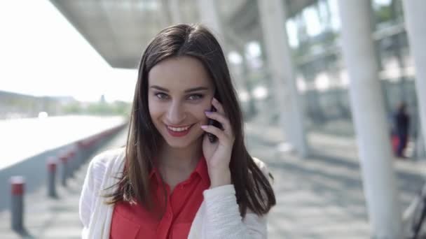 Mulher de camisa vermelha fala ao telefone andando com uma mala — Vídeo de Stock