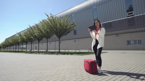 Жінка читає щось на планшеті, стоячи з червоною валізою — стокове відео